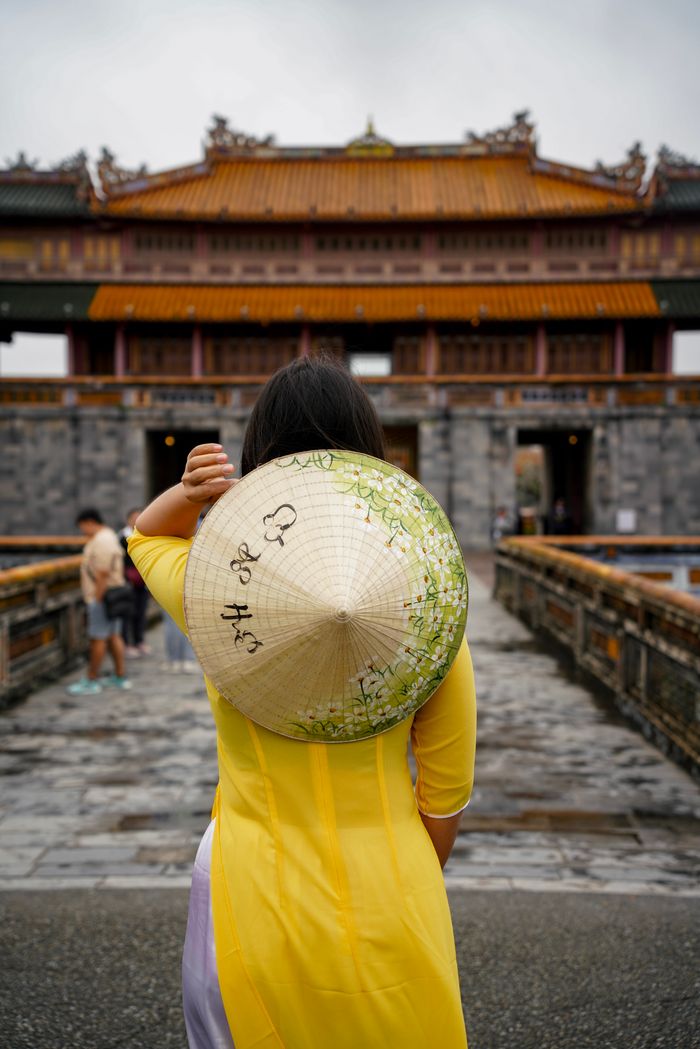 A woman in traditional Vietnamese dress and hat stands before an ancient temple.