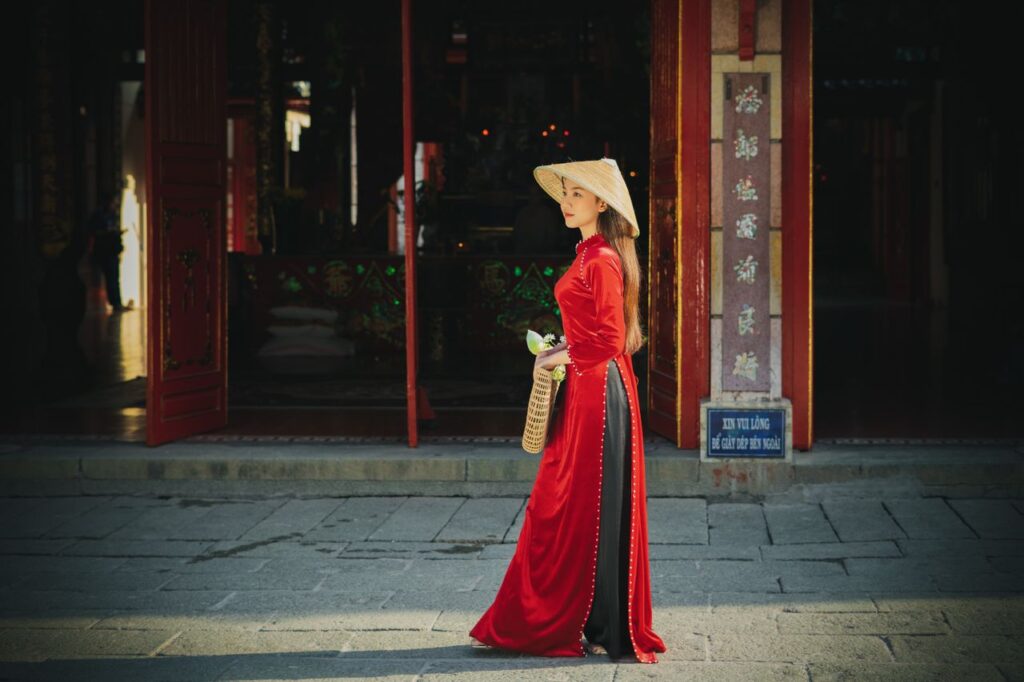 An Asian woman in a red dress and conical hat walking outdoors, embodying cultural elegance.