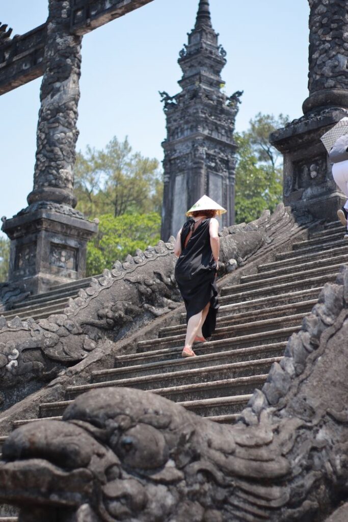 A woman in a black dress climbs ancient stone stairs wearing a conical hat.