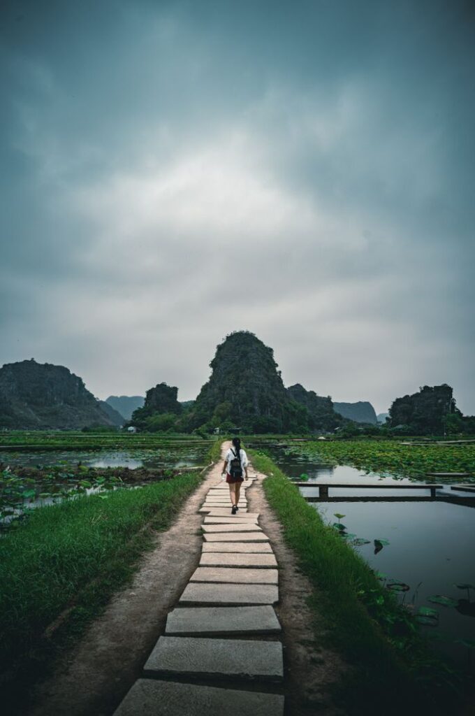 Traveler walking along a stone path through lush landscape of Ninh Bình, Vietnam under a moody sky.