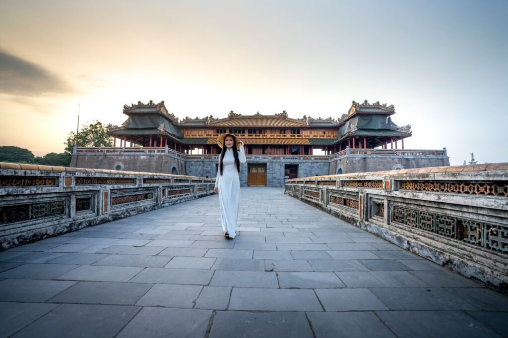 Full body of female in white dress and hat strolling on walkway near aged building and stone fence under cloudless sky in twilight